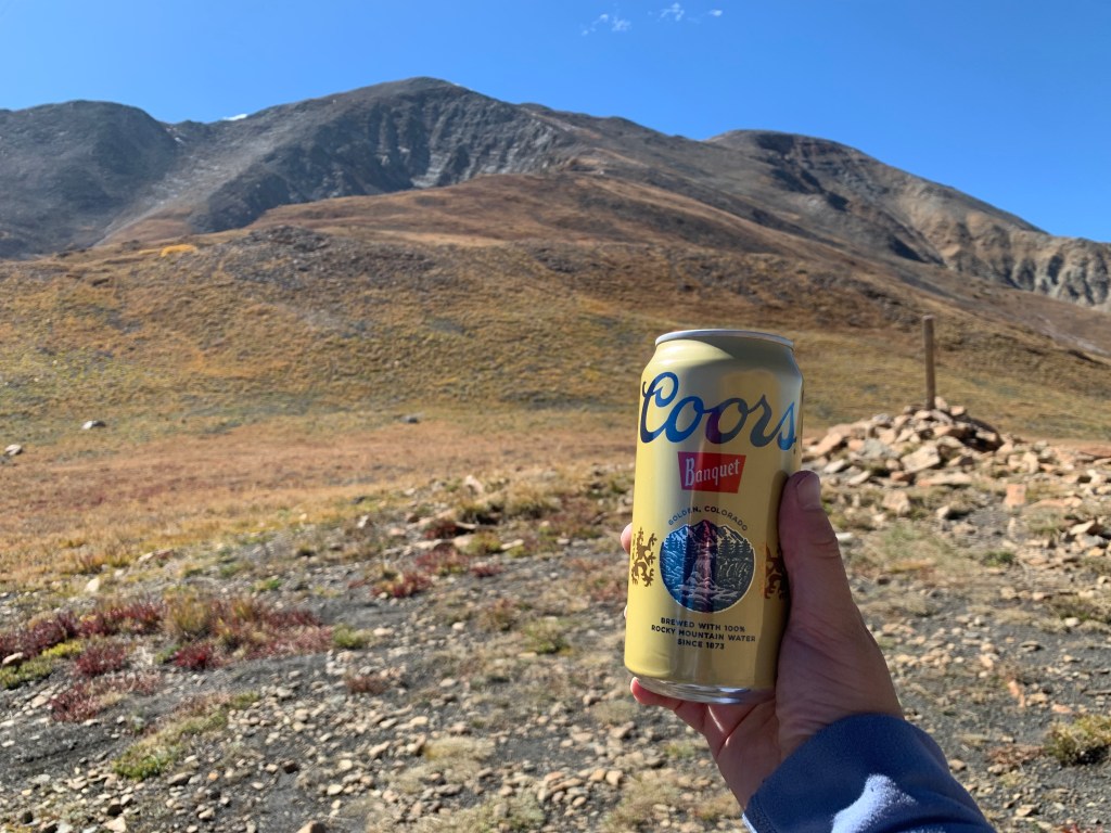 A light-skinned right hand holds up a can of Coors Banquet in front of a high alpine peak with reddish-brown autumnal tundra grasses and rocks below a blue sky.